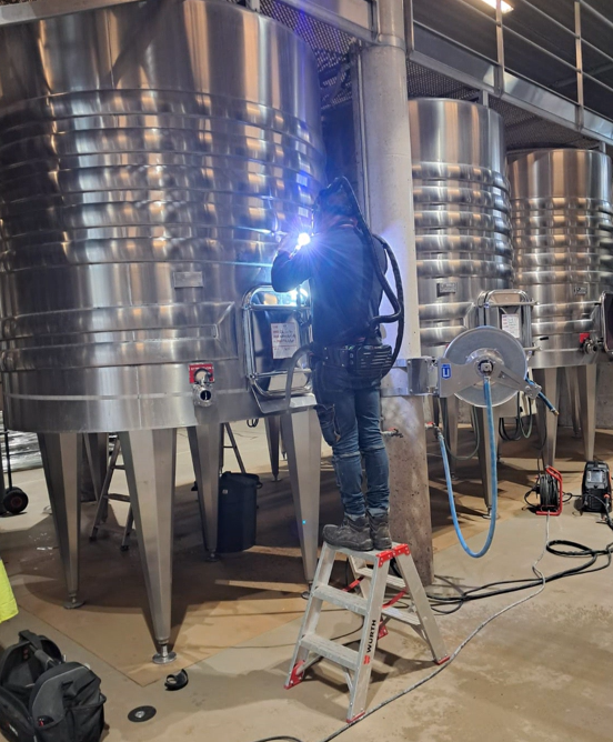 Un ouvrier soude un réservoir sur un escabeau dans une cave à vin, produisant une lumière vive.