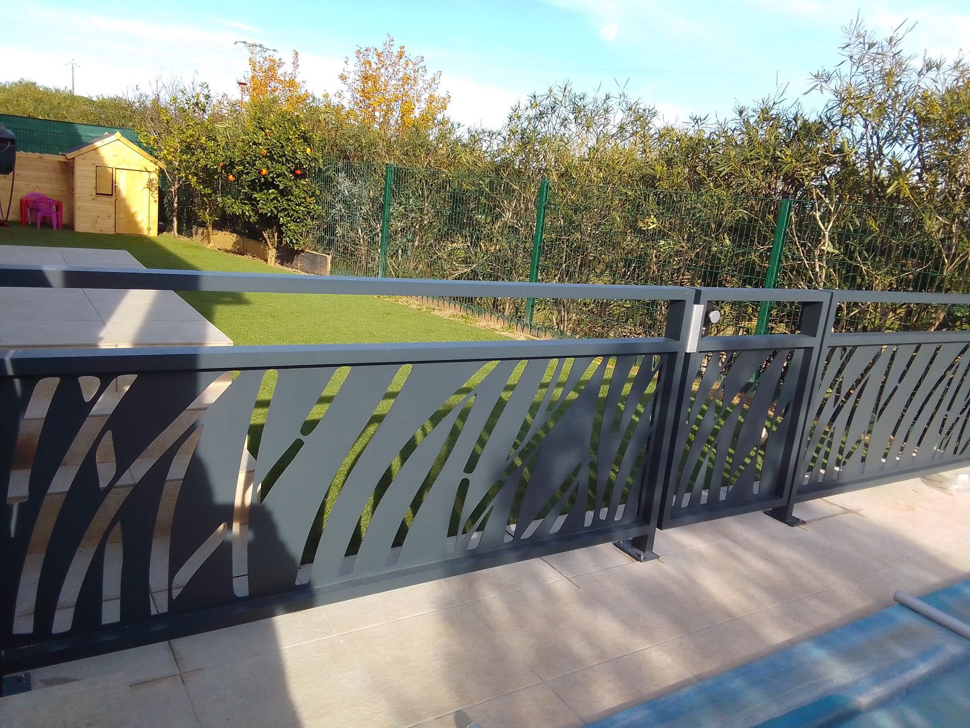 Clôture en métal gris avec un motif découpé en forme de feuille, devant une cour verte et un petit bâtiment jaune.