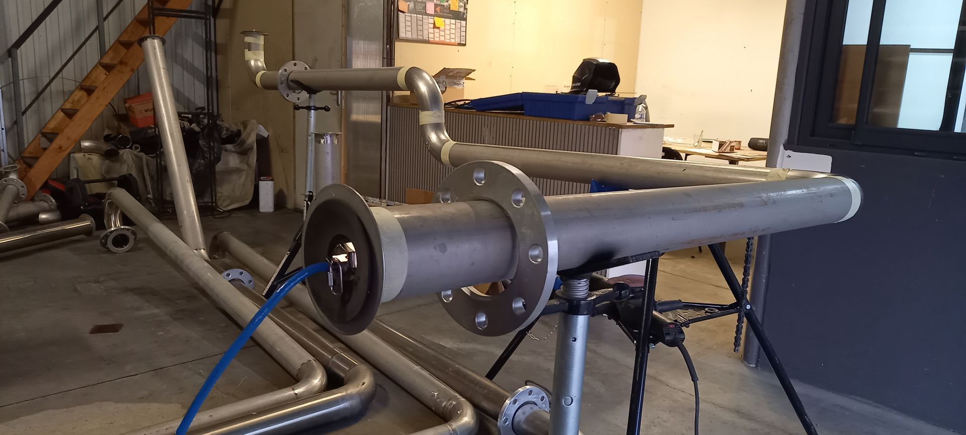 Un tuyau avec des brides et un câblage est monté sur un trépied dans une pièce.