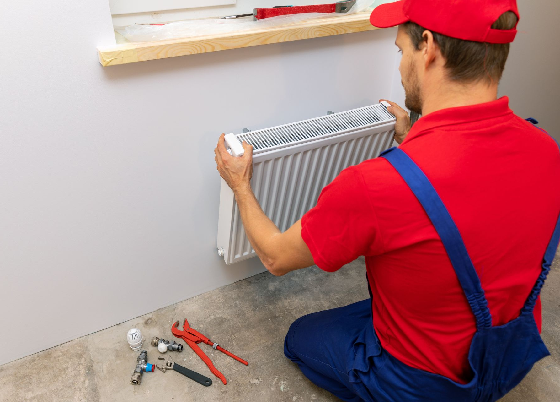 Un ouvrier vêtu d'une chemise et d'une salopette installe un radiateur en métal sur un mur, tandis que des outils sont posés à proximité, sur le sol.