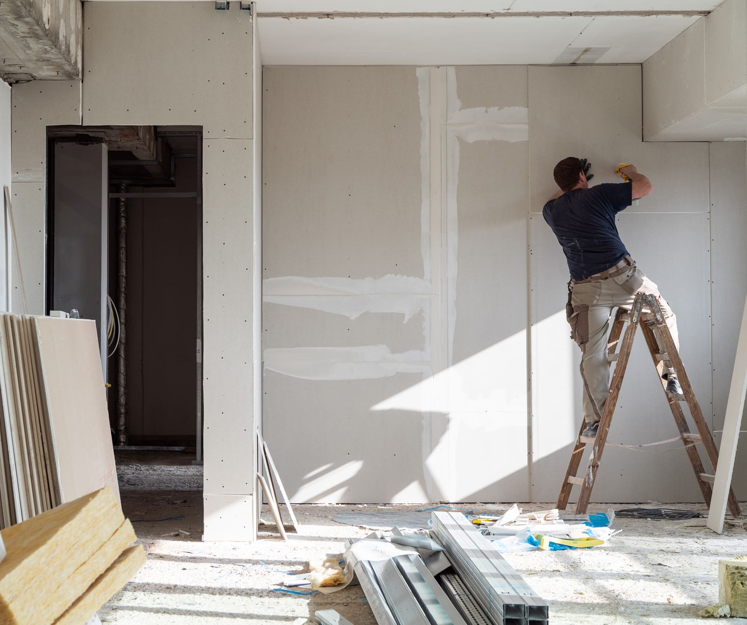 Une personne, juchée sur une échelle, applique de l'enduit à joints sur les raccords d'une cloison sèche nouvellement installée dans une pièce non finie.