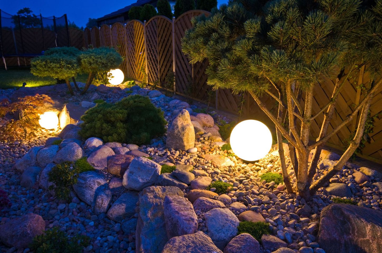 Au crépuscule, des lampes sphériques lumineuses éclairent des rochers, des arbustes et une clôture en bois dans un jardin.
