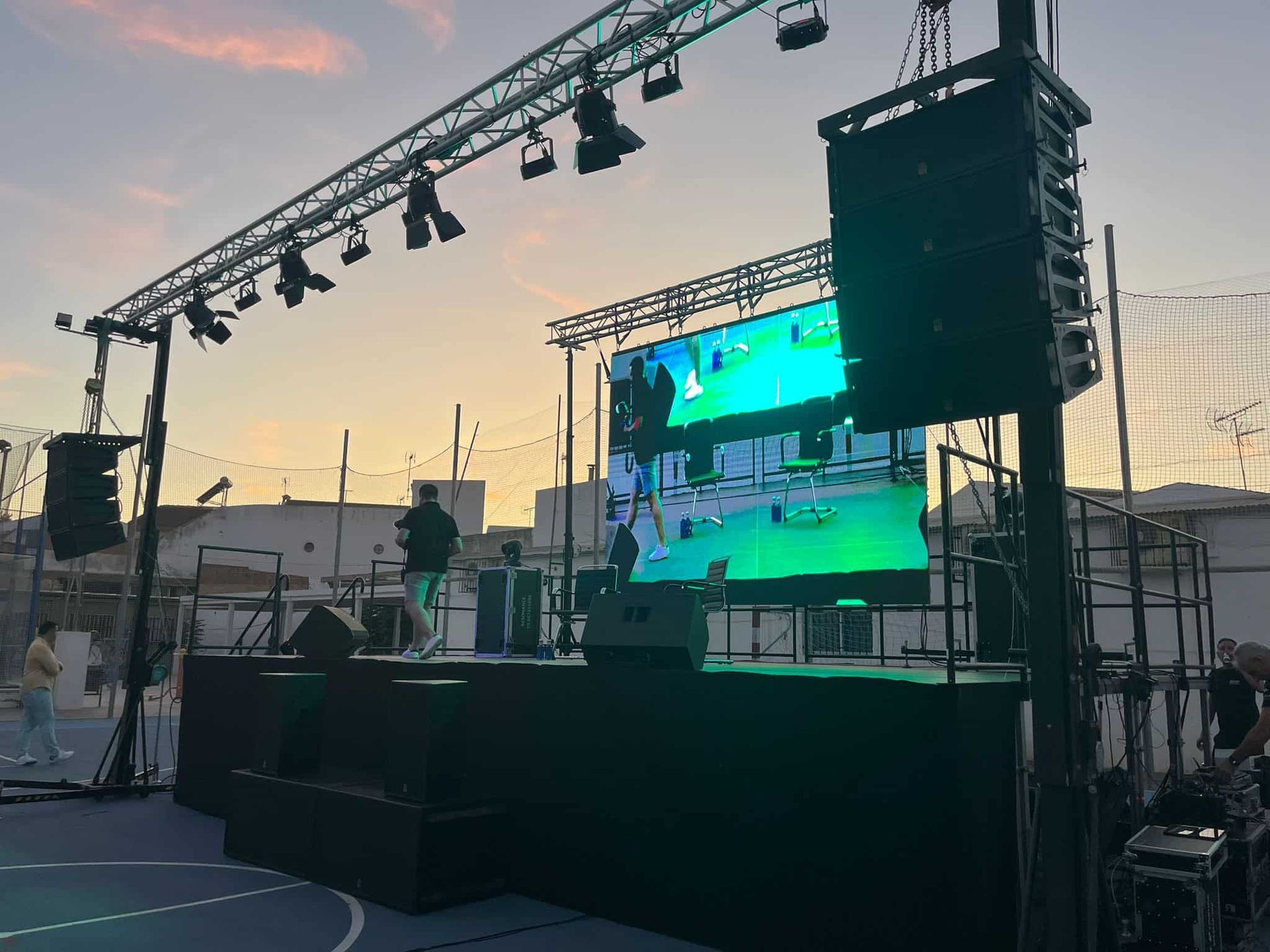 Escenario al aire libre con equipo de iluminación y pantalla de vídeo, una persona en el escenario actuando durante el anochecer.