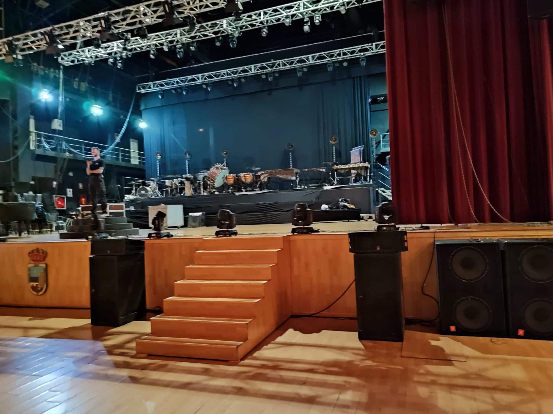 Escenario con instrumentos, iluminación y equipo de sonido. Una persona está de pie en el escenario. Cortinas rojas a la derecha.