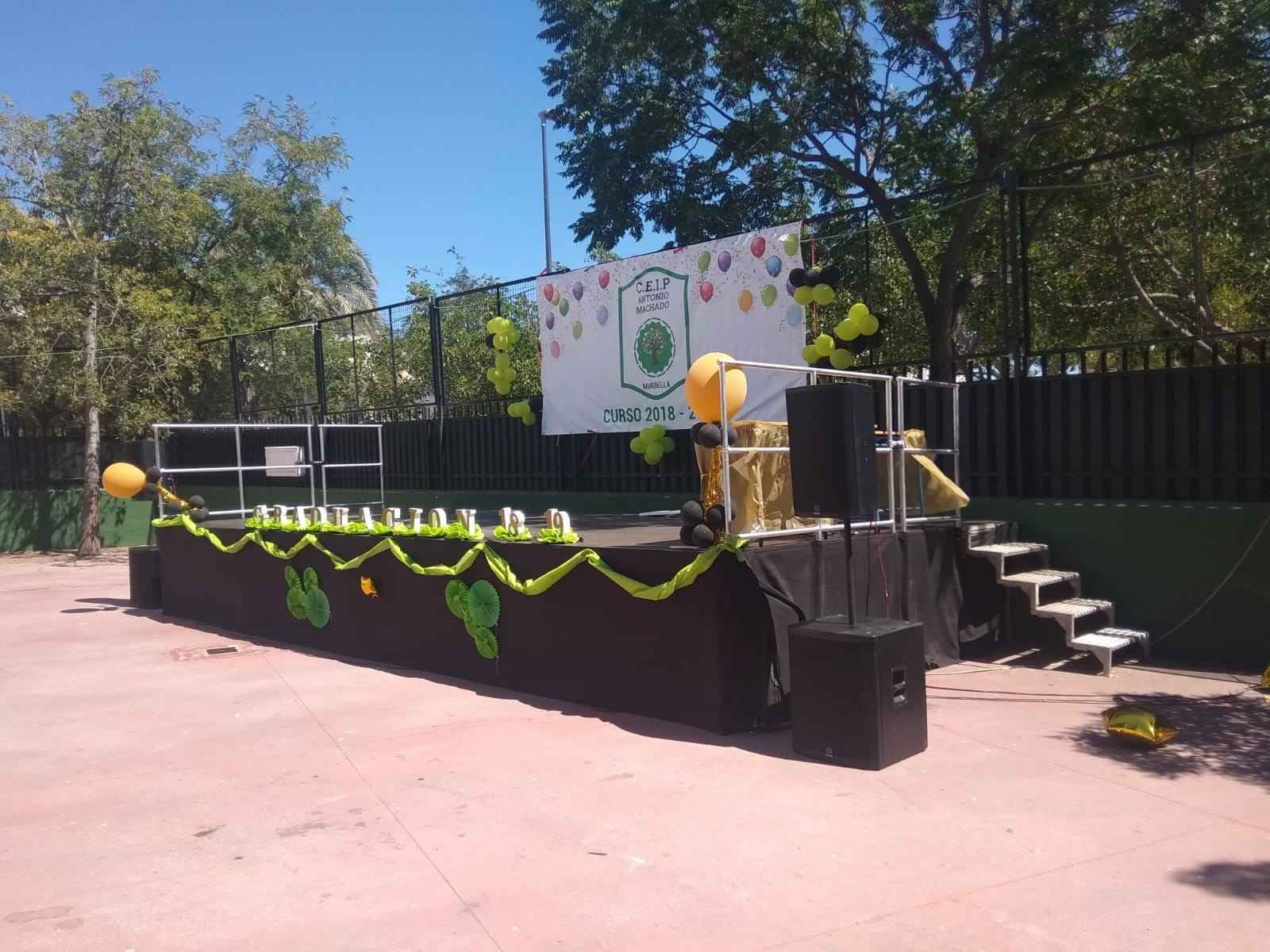 Escenario al aire libre decorado con globos, guirnaldas y letrero. Al fondo, suelo rojo, árboles y una valla.