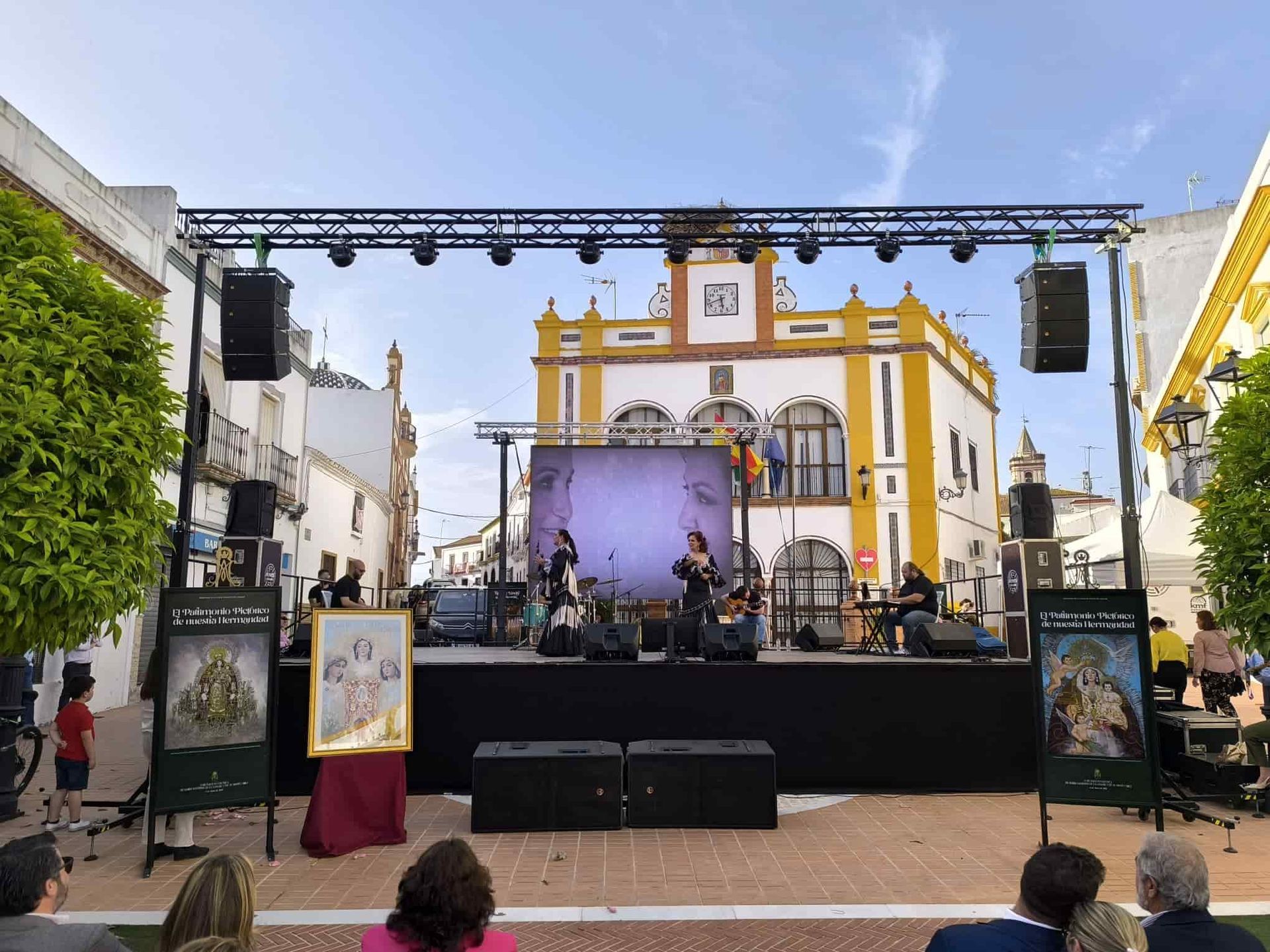 Banda tocando en un escenario frente a un edificio amarillo y blanco en una plaza de la ciudad.