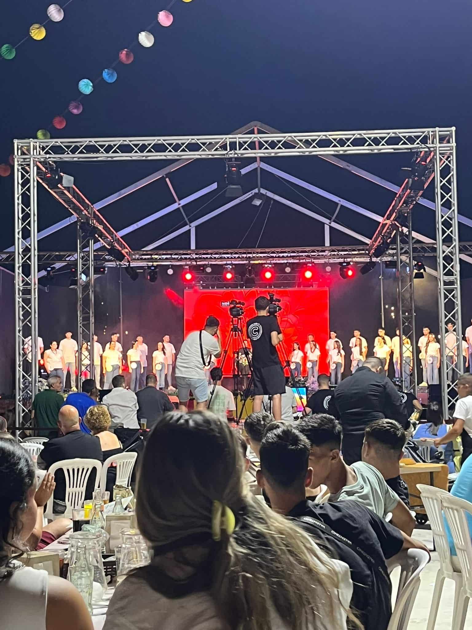 Personas viendo una actuación en un escenario en un evento al aire libre, con mesas y luces de cadena.