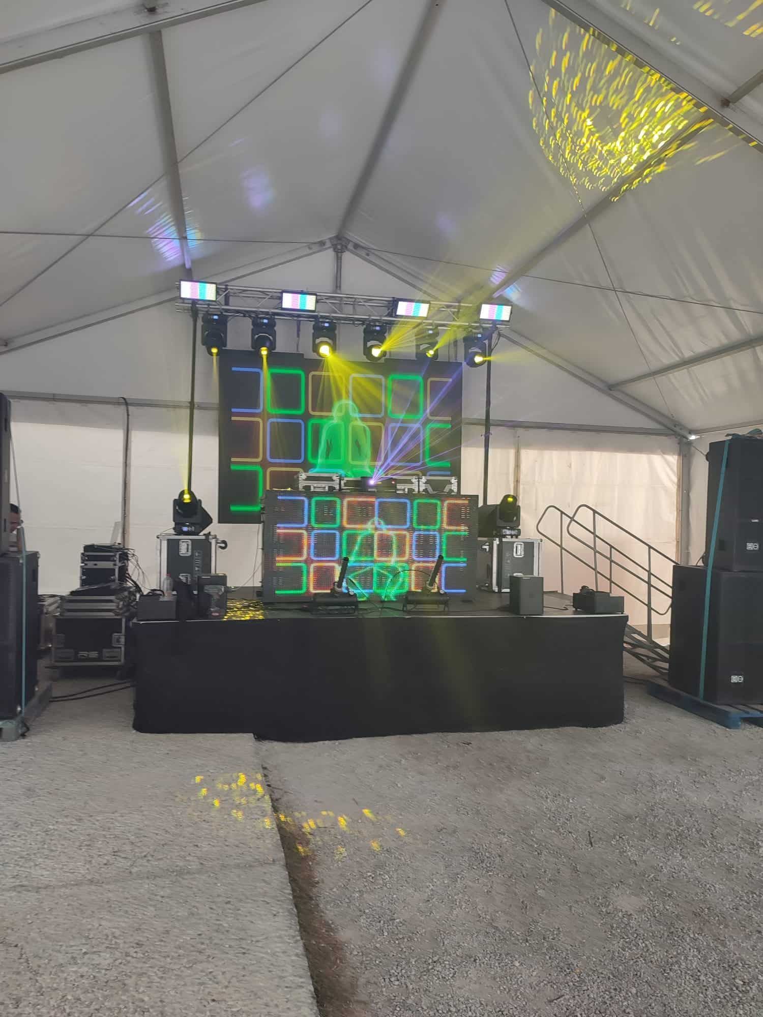 Montaje de DJ en escenario bajo carpa, con luces y equipo de sonido.
