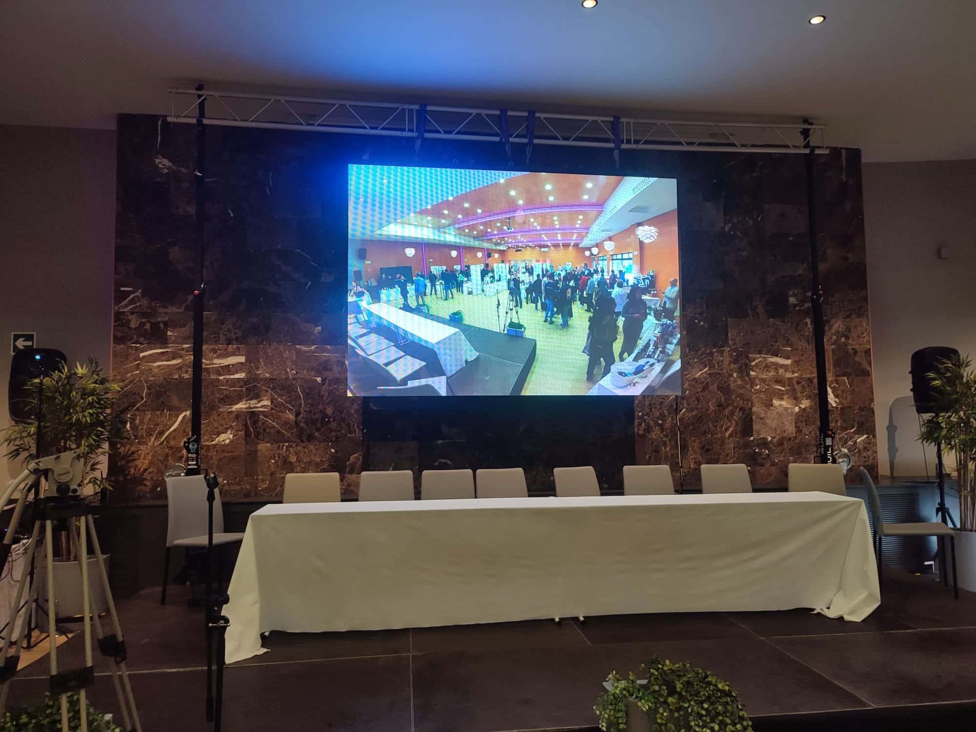 Una gran pantalla de vídeo muestra el evento; frente a ella hay una mesa con sillas.