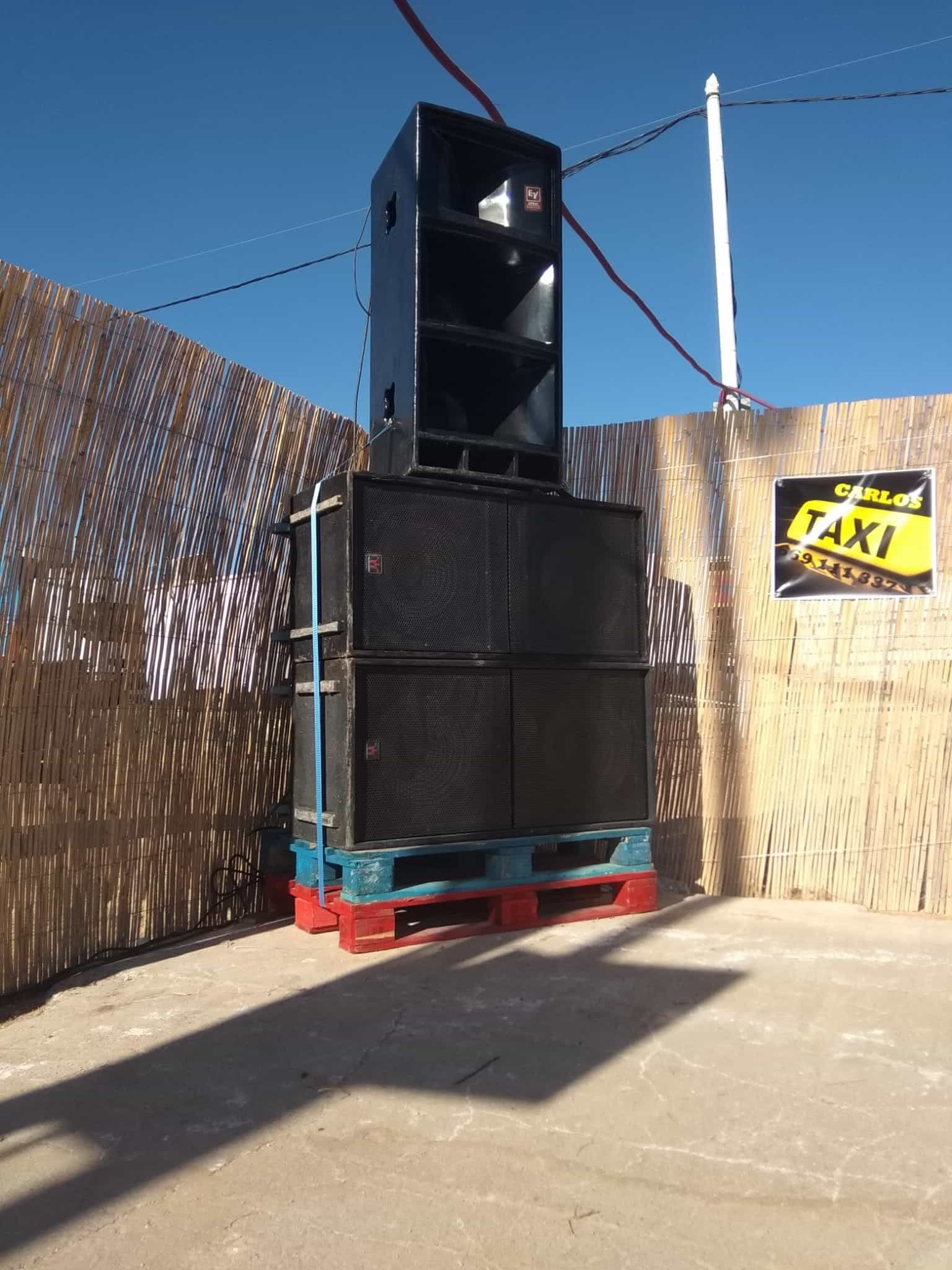 Sistema de sonido exterior: altavoces apilados, cajas negras sobre palés, contra una pared de metal corrugado, señal de taxi.