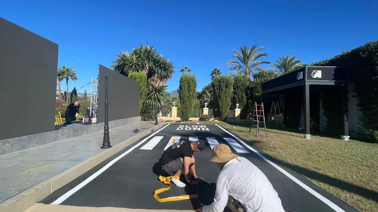 Dos personas trabajan en una carretera pintada con paso de peatones, rodeadas de vegetación y edificios, bajo un cielo azul.
