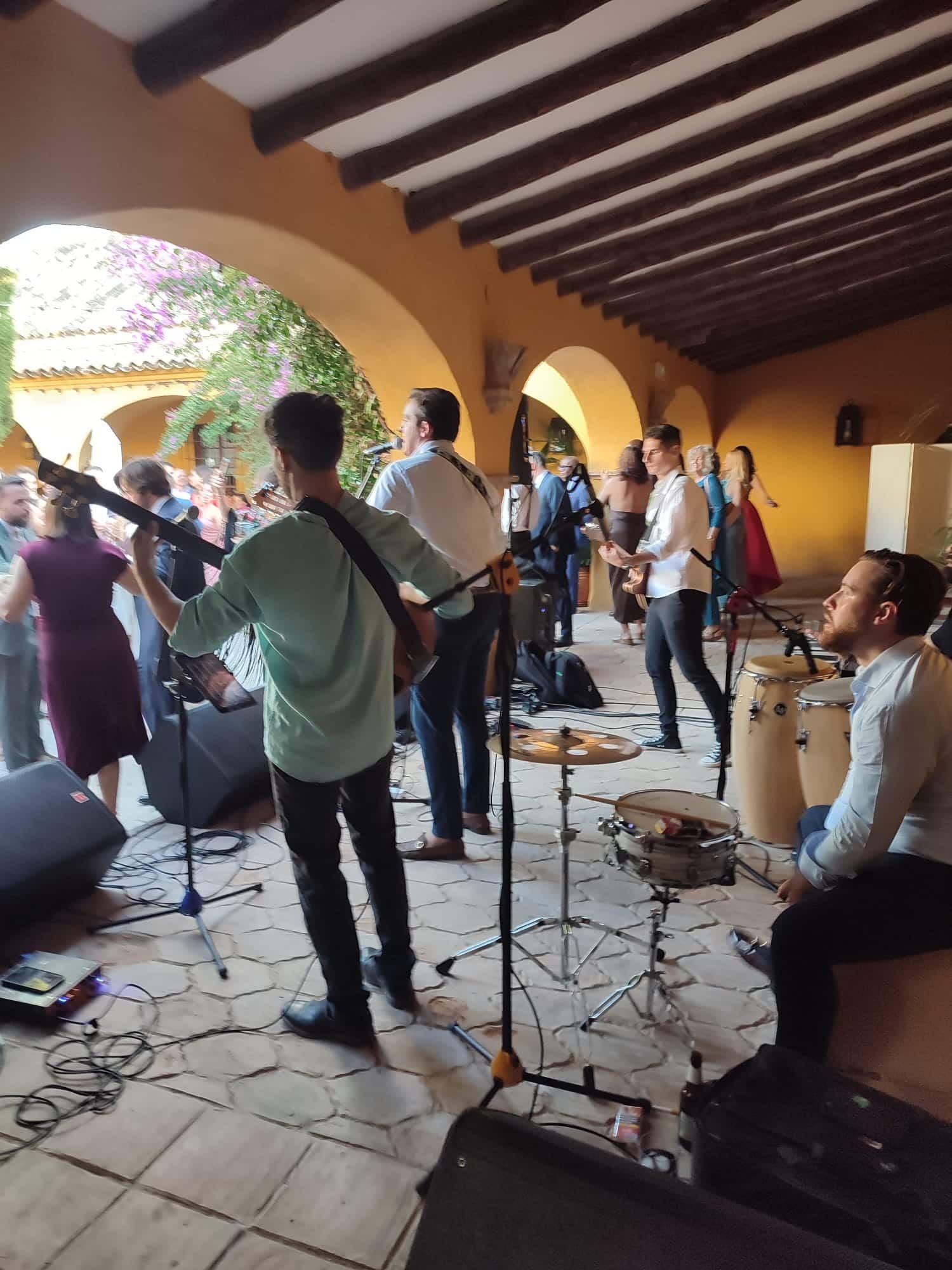 Banda tocando música en vivo al aire libre bajo un techo abovedado. Gente al fondo.