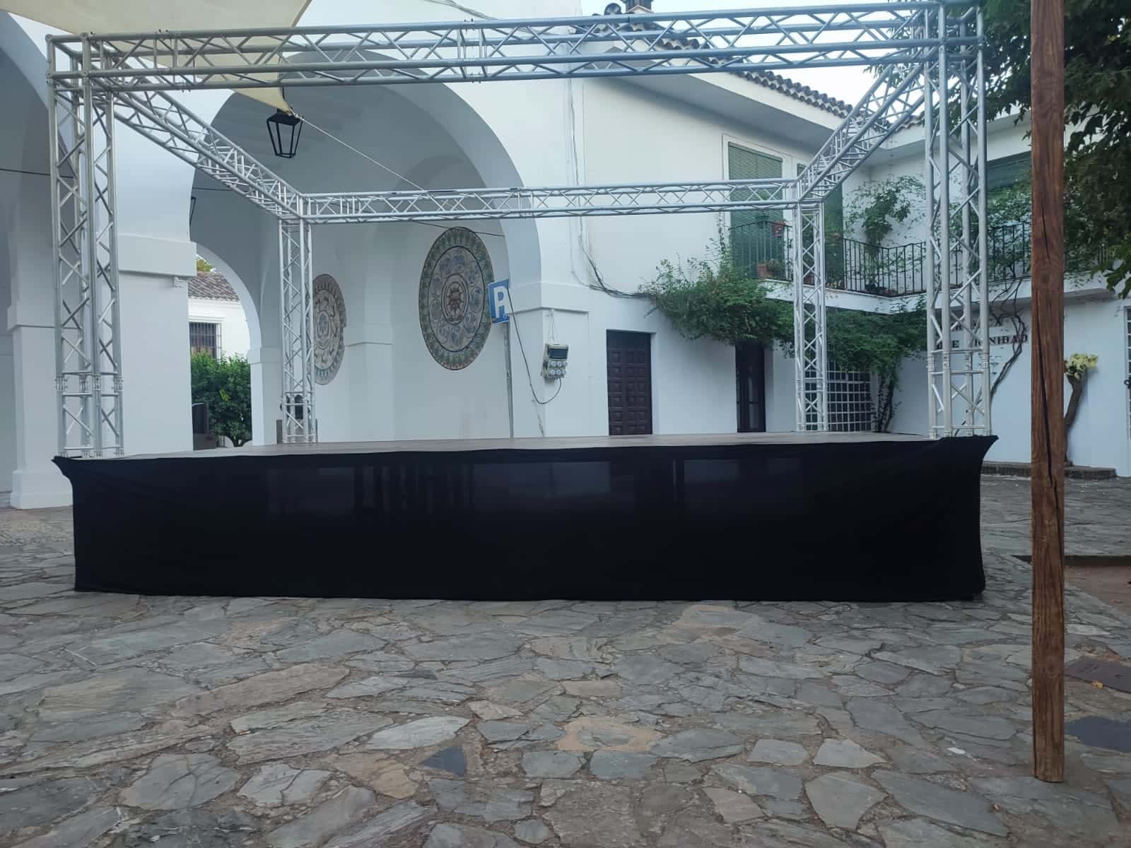 Escenario con faldón negro y estructura metálica, situado sobre suelo adoquinado frente a edificio blanco.