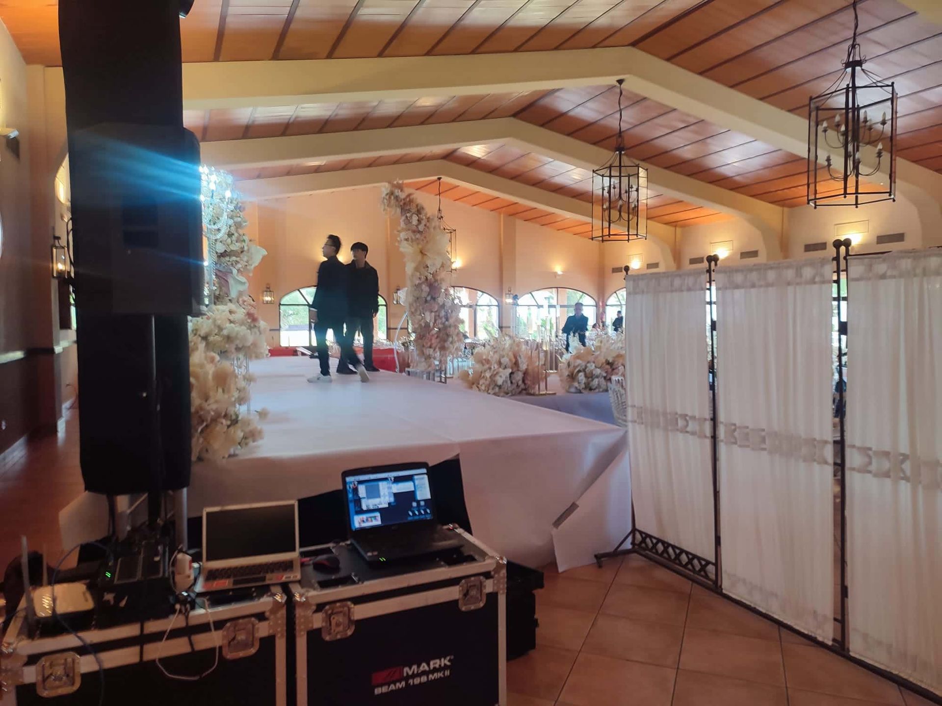 Montaje de recepción de boda con escenario, equipo de DJ y decoración. Dos personas de pie en el escenario.