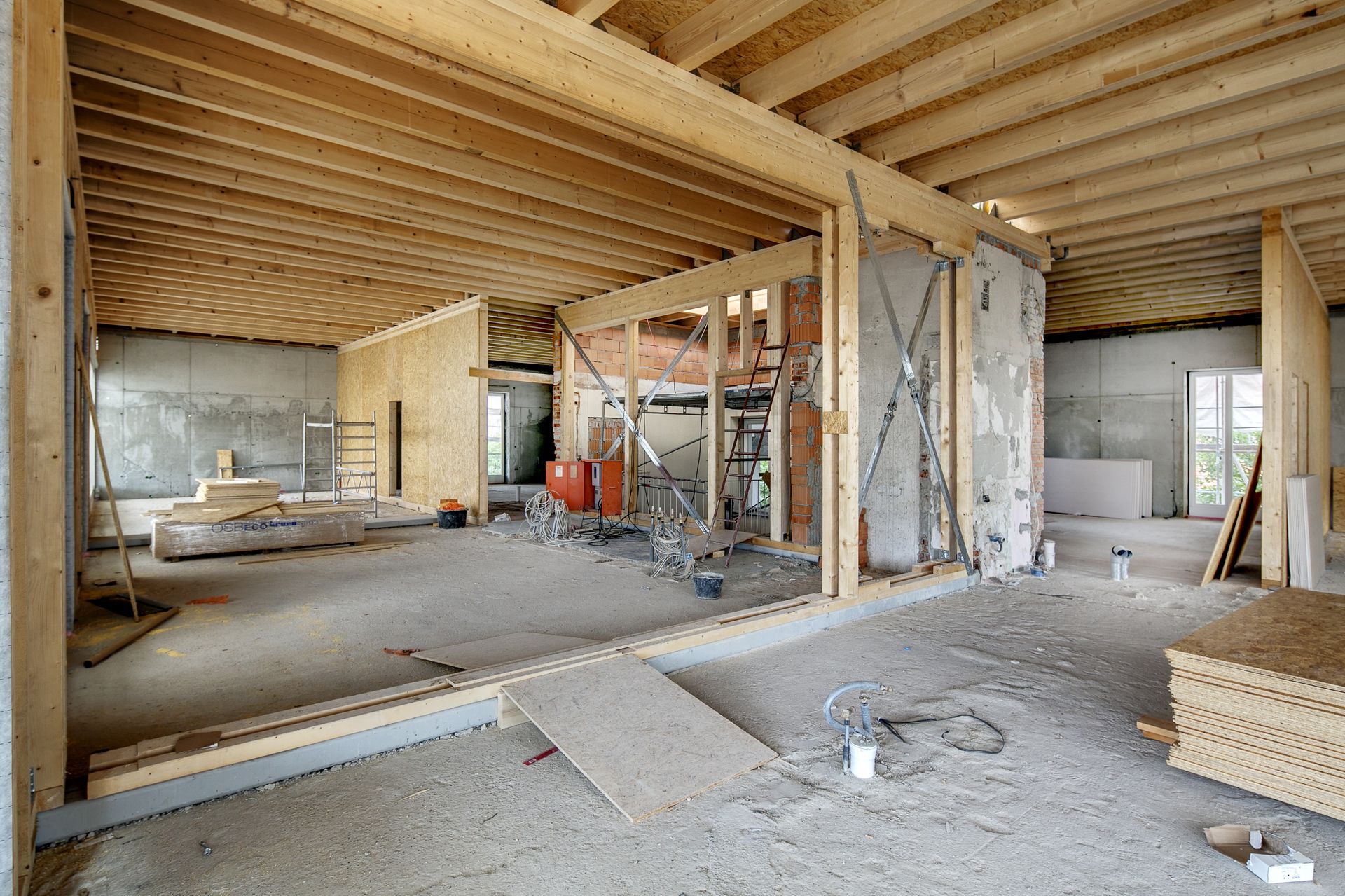 Innenansicht eines im Bau befindlichen Gebäudes mit freiliegenden Holzbalken, Betonwänden und Baumaterialien.