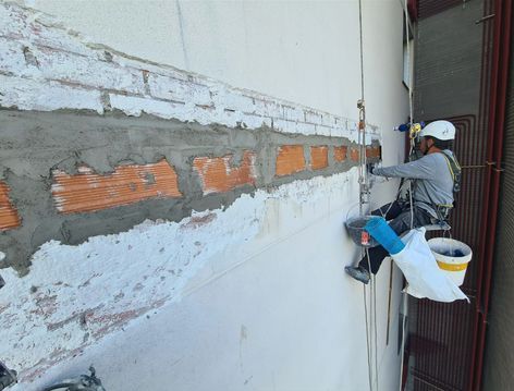 Un trabajador repara una grieta en la pared exterior junto a una ventana, utilizando un cubo y herramientas.