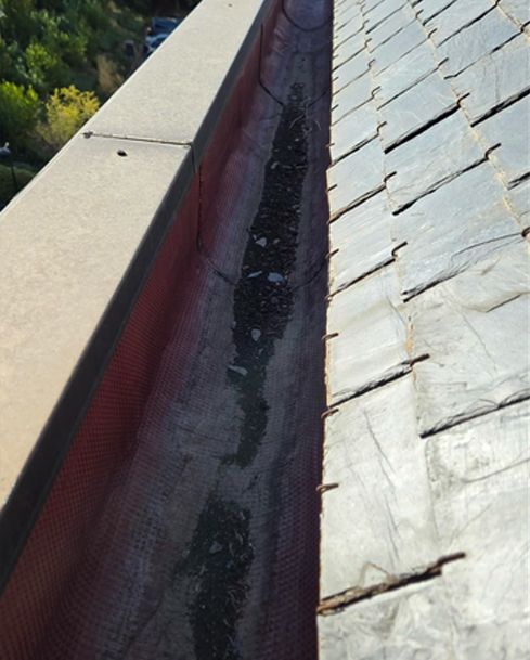 Primer plano de un canalón de tejado con escombros negros y una tira de sellado roja entre tejas grises.