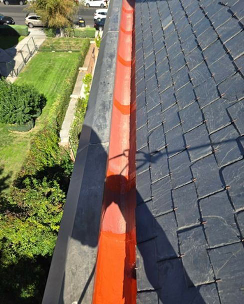 Canalón naranja sobre techo de tejas oscuras junto a un jardín paisajístico y entrada para vehículos.