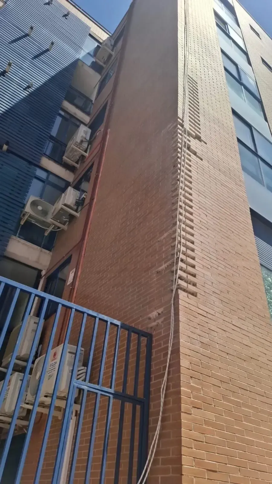 Fachada de un edificio alto de ladrillo junto a una estructura con marco azul y una puerta de metal, vista desde abajo.