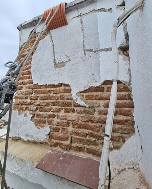 Pared de yeso agrietada que deja al descubierto el ladrillo rojo y una tubería blanca inclinada con escombros en la base.