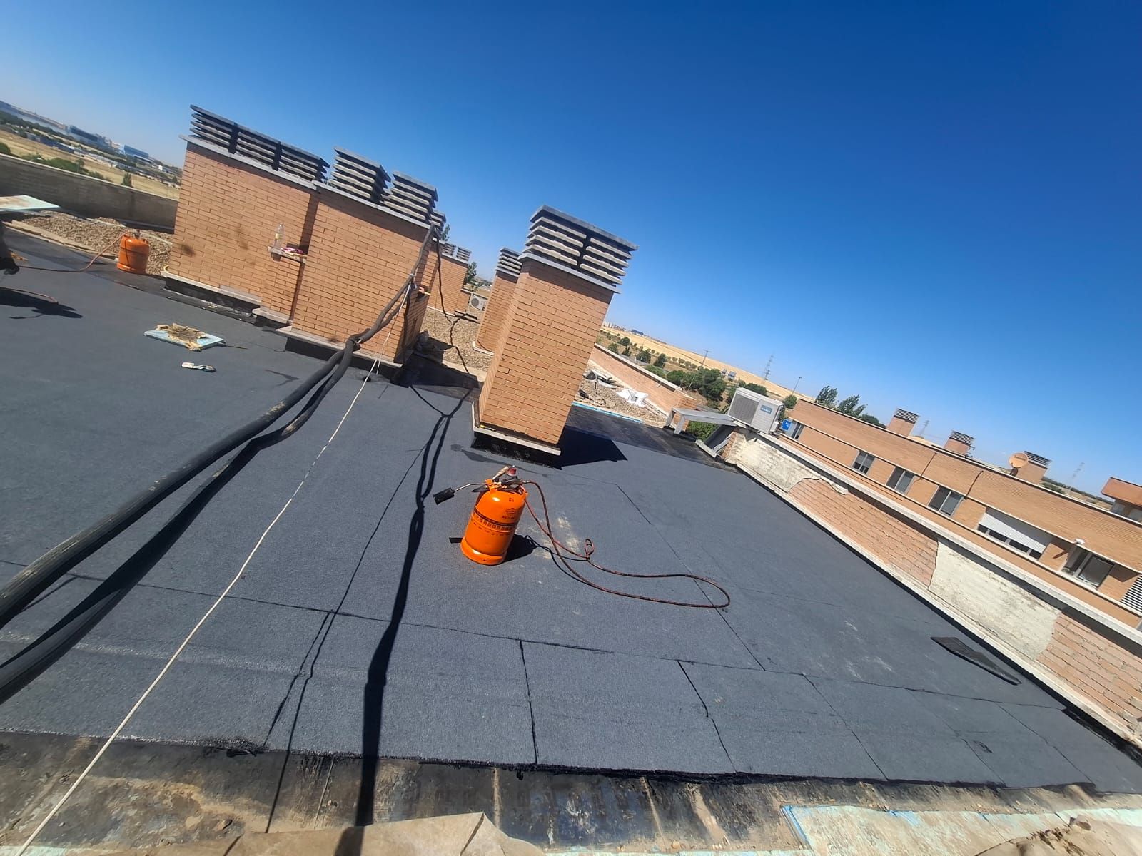 Azotea con membrana negra, tubo de ventilación naranja y estructuras de ladrillo para servicios públicos bajo un cielo azul claro.