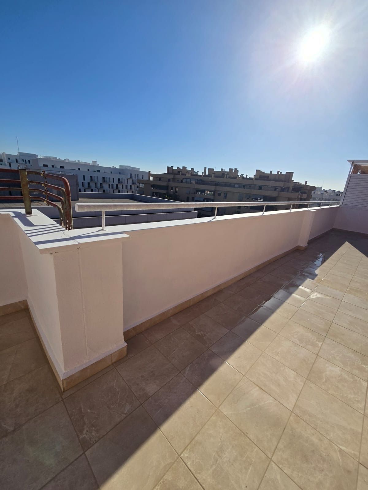 Terraza soleada en la azotea con suelo de baldosas beige, pared baja blanca y edificios de la ciudad a lo lejos.