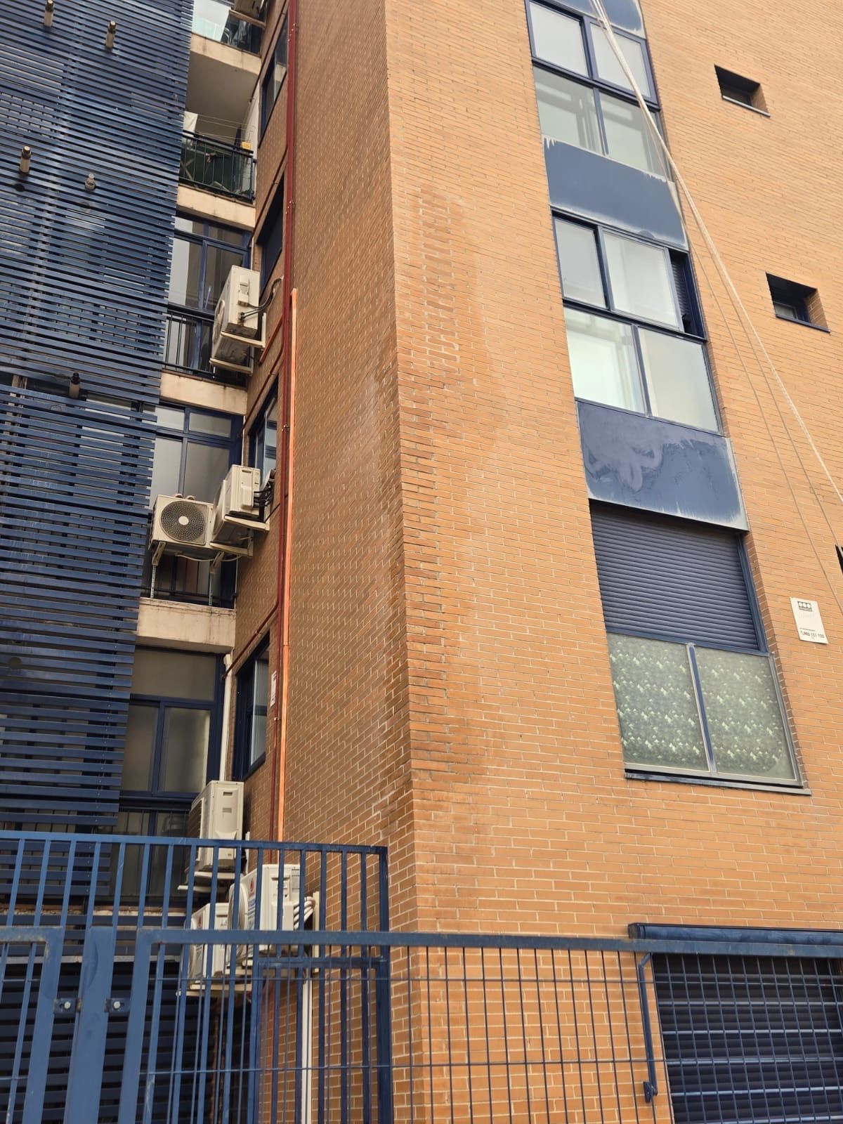 Edificio de apartamentos alto de ladrillo con paneles metálicos azules y balcones detrás de una valla negra.