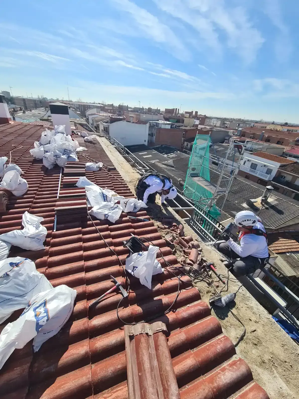 Trabajadores reparando un tejado de tejas rojas bajo un cielo azul, con herramientas y escombros esparcidos por la azotea.