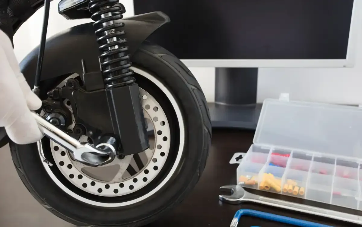 Una persona con guantes blancos repara una rueda de scooter eléctrico con una llave inglesa cerca de una caja de herramientas y un monitor de computadora.