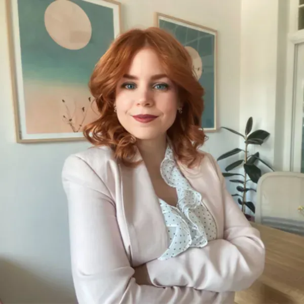 Mujer pelirroja sonriente con los brazos cruzados en una habitación luminosa, vestida con un blazer claro
