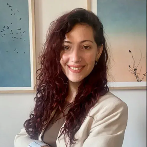 Mujer sonriente con cabello oscuro y rizado, vestida con una blusa blanca, sentada en el interior