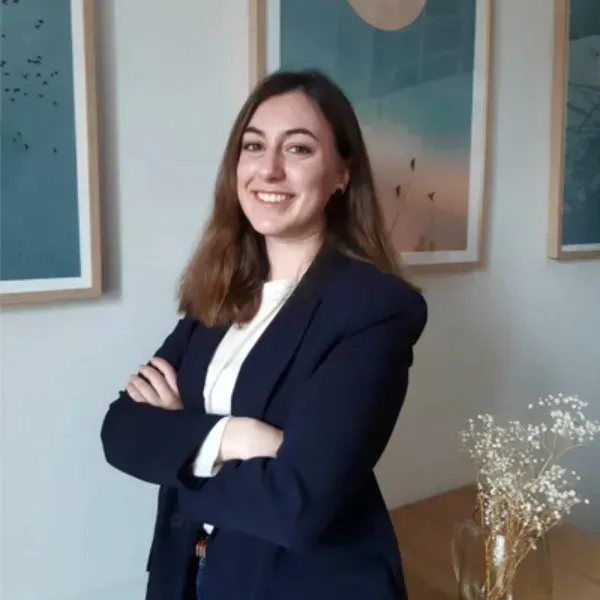 Persona sonriente con un blazer azul marino y los brazos cruzados, de pie en el interior de una casa junto a un cuadro