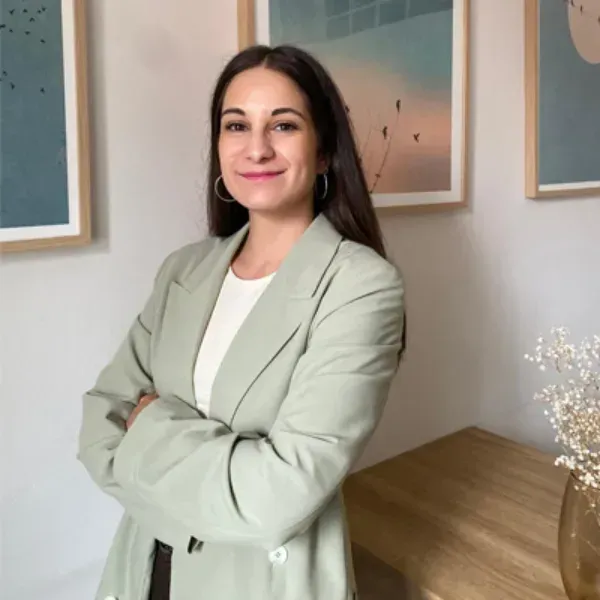 Una persona con una chaqueta verde claro permanece de pie con los brazos cruzados en una oficina moderna, sonriendo.