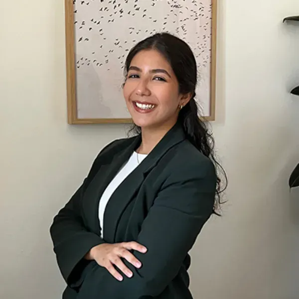 Mujer sonriente con chaqueta oscura y brazos cruzados, de pie en el interior de una casa junto a un cuadro enmarcado.