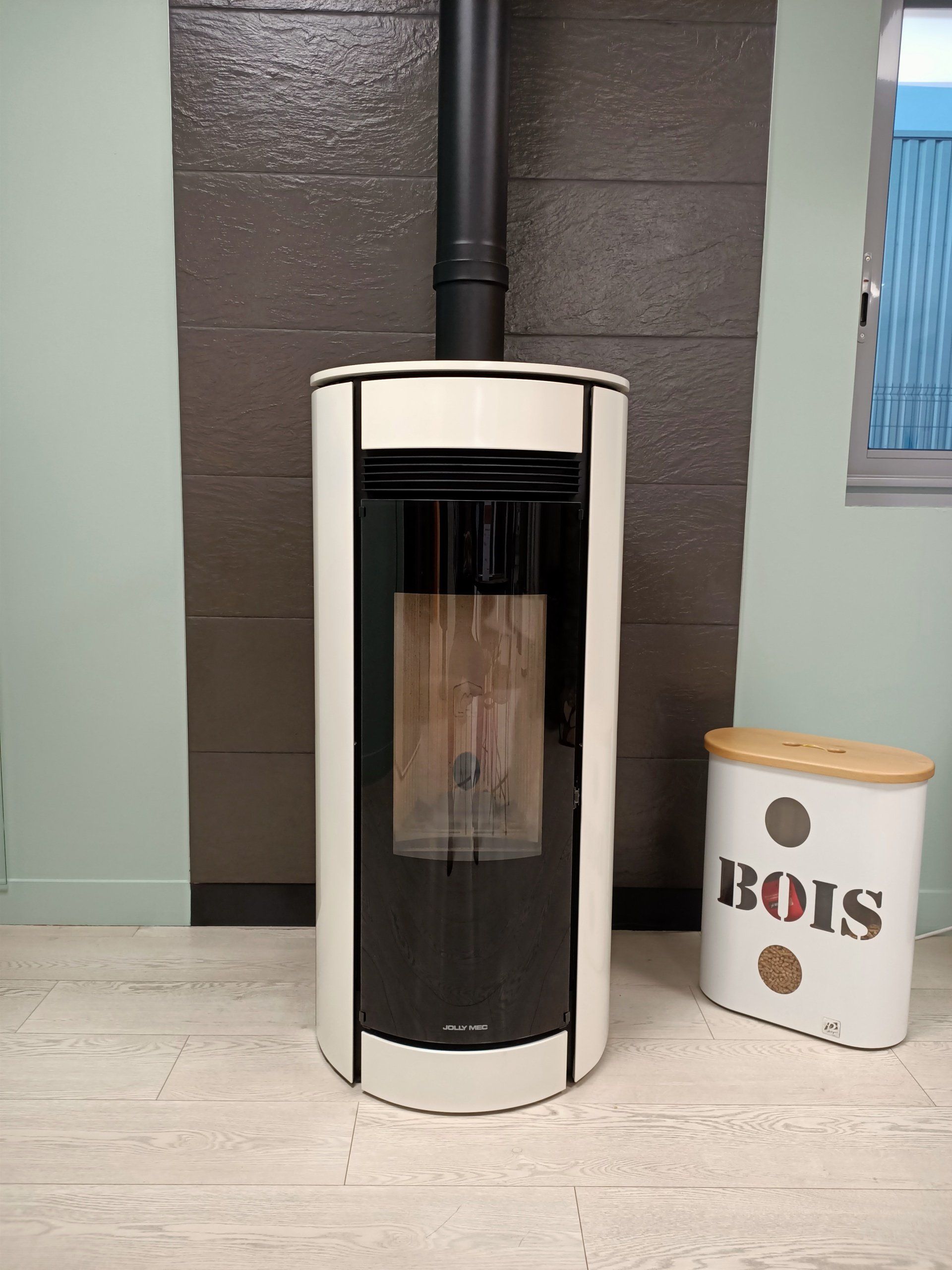 Poêle à bois blanc et noir avec une cheminée noire, posé sur un sol clair contre un mur brun gris.