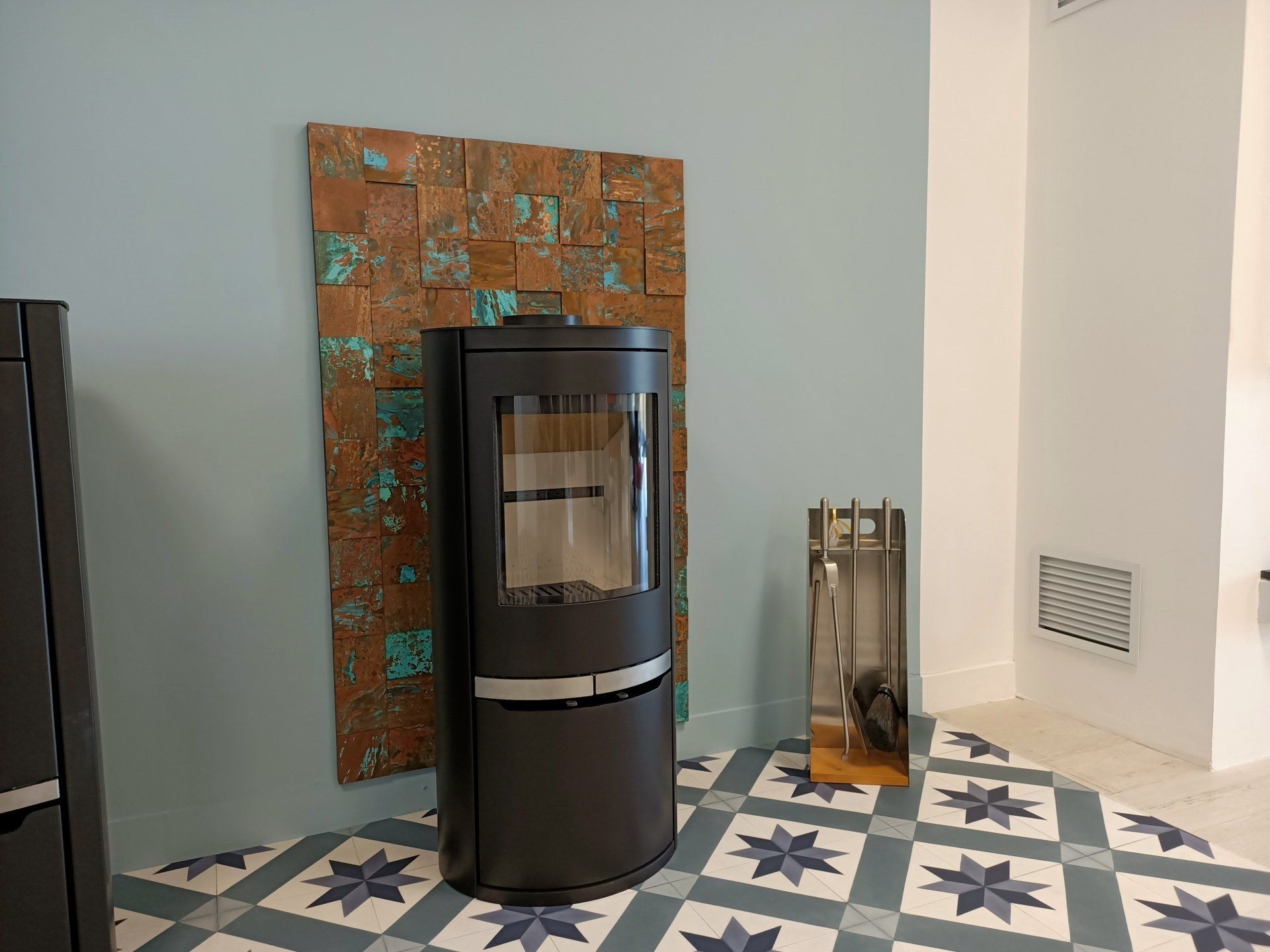 Poêle noir contre un mur bleu avec des carreaux de cuivre, sur un sol à motifs