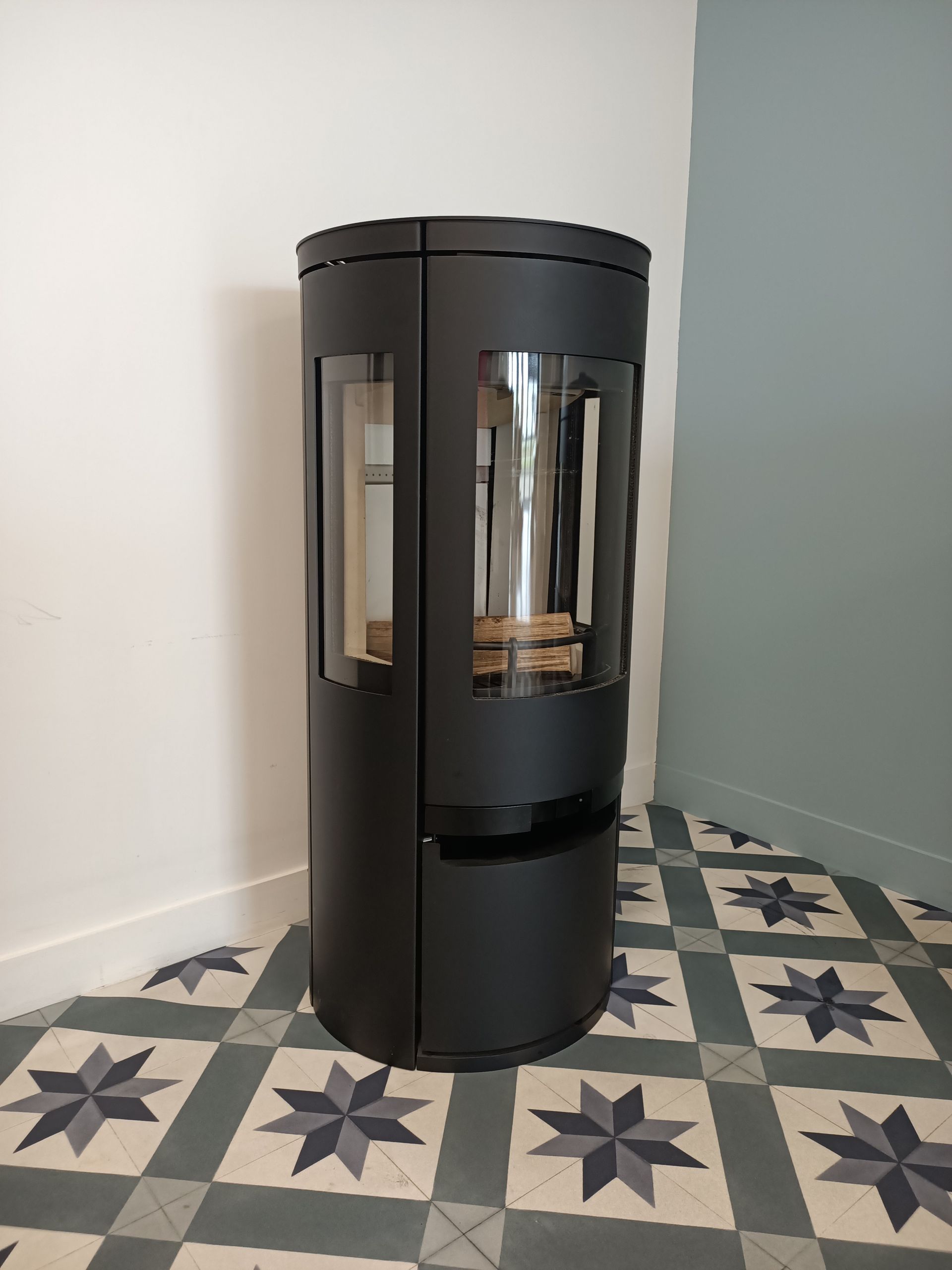 Poêle à bois cylindrique noir dans un coin, avec panneaux vitrés. Carrelage au sol