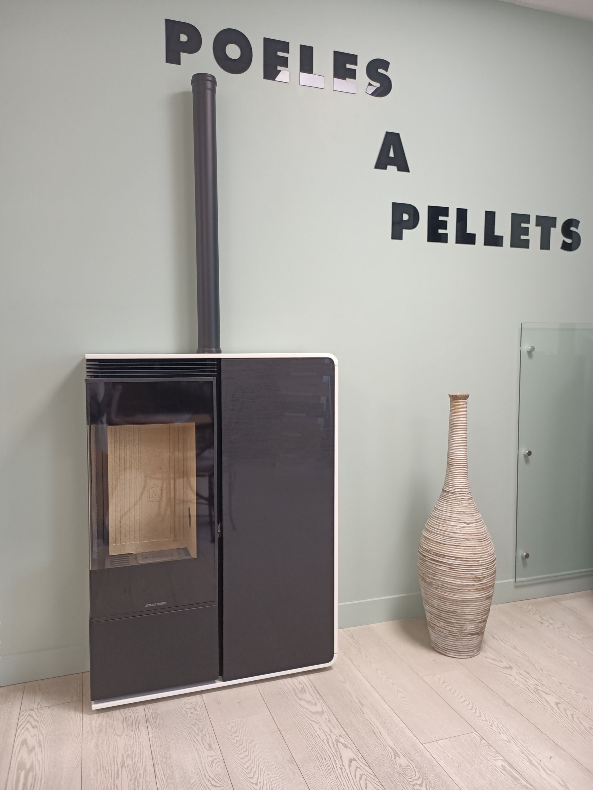 Un poêle à granulés noir et blanc, avec une cheminée contre un mur vert pâle. Un vase décoratif trône à côté.