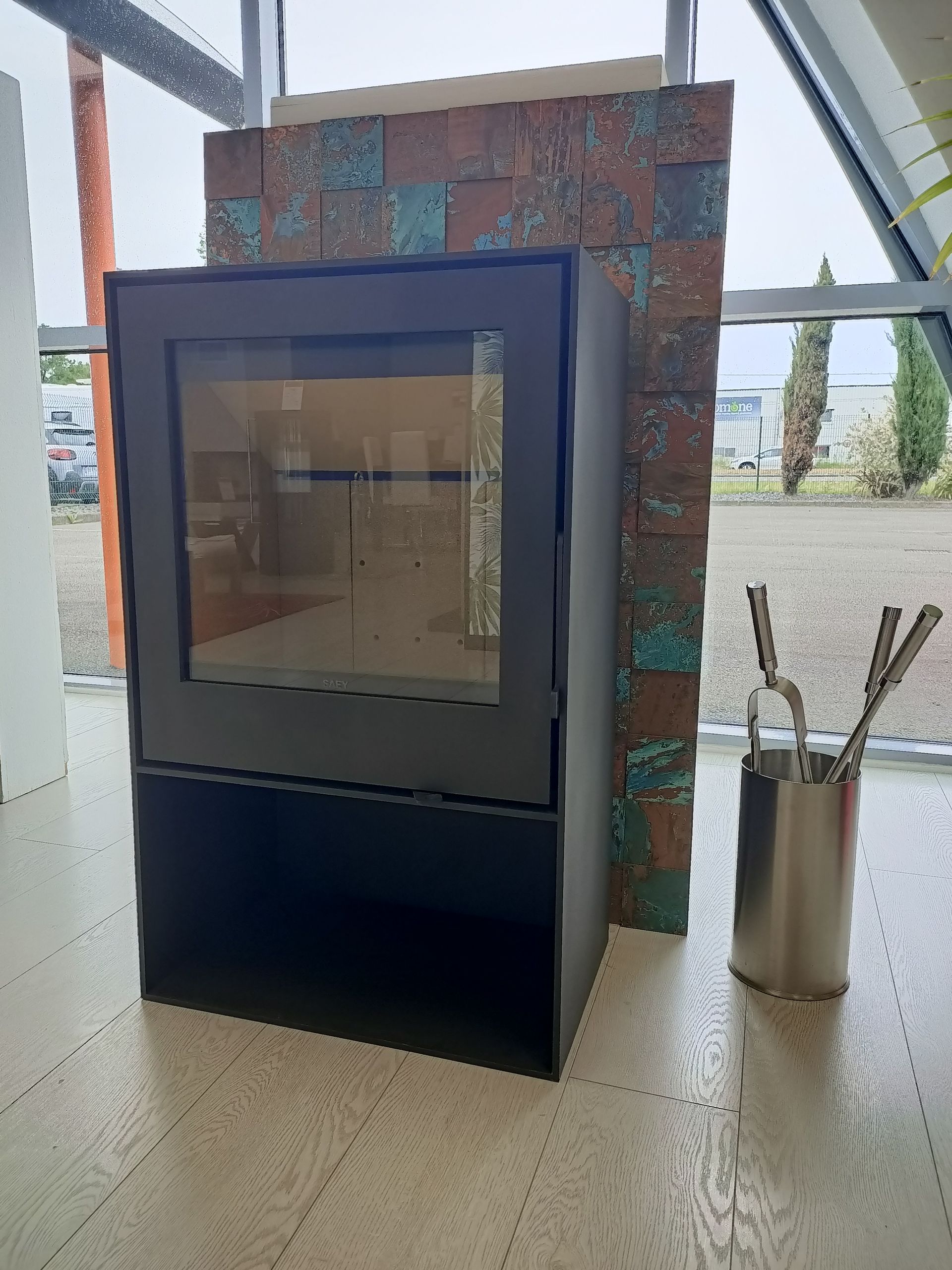 Cheminée noire avec façade en verre, à côté d'un mur texturé et d'un porte-outil en métal, fenêtre en arrière-plan.