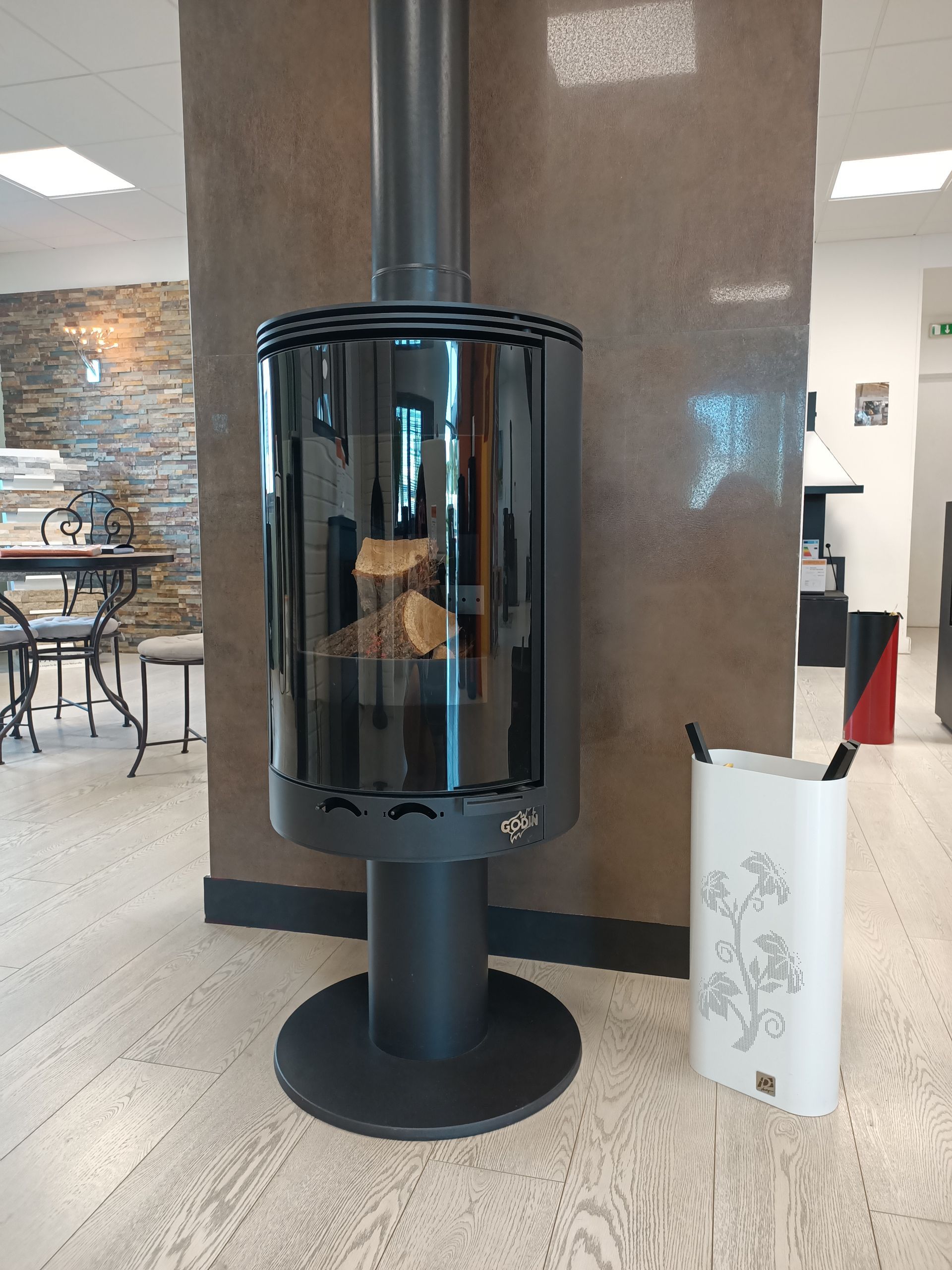 Cheminée cylindrique noire avec panneaux en verre, sur une base noire, avec du bois de chauffage à l'intérieur