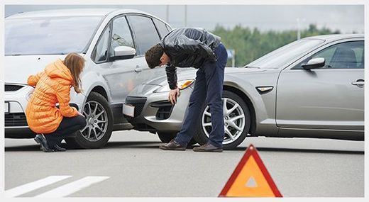 Dos personas inspeccionan un coche dañado tras una colisión en la carretera, con un triángulo de advertencia delante.