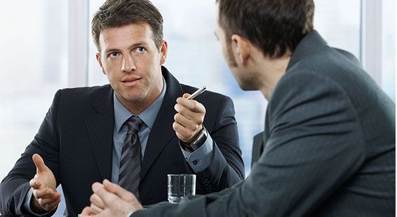Dos hombres de negocios con traje conversan sentados a una mesa; uno gesticula mientras el otro escucha atentamente.