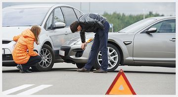 Dos personas cambian un neumático de un coche plateado tras una avería en la carretera, con un triángulo de advertencia cerca.