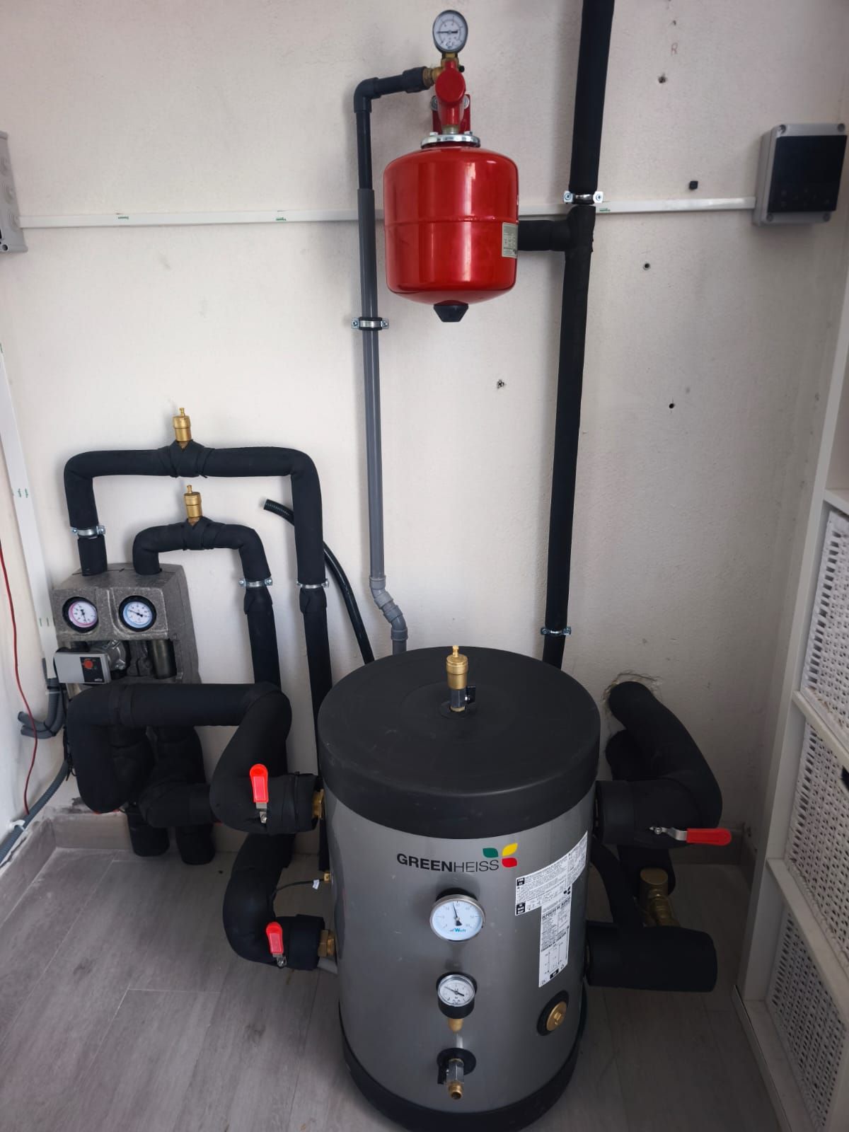 Cuarto de calderas con un depósito de agua caliente gris, un tanque de expansión rojo, tuberías negras y manómetros, con paredes blancas