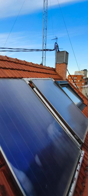 Paneles solares en un tejado de tejas rojas bajo un cielo azul, con una antena de televisión al fondo.