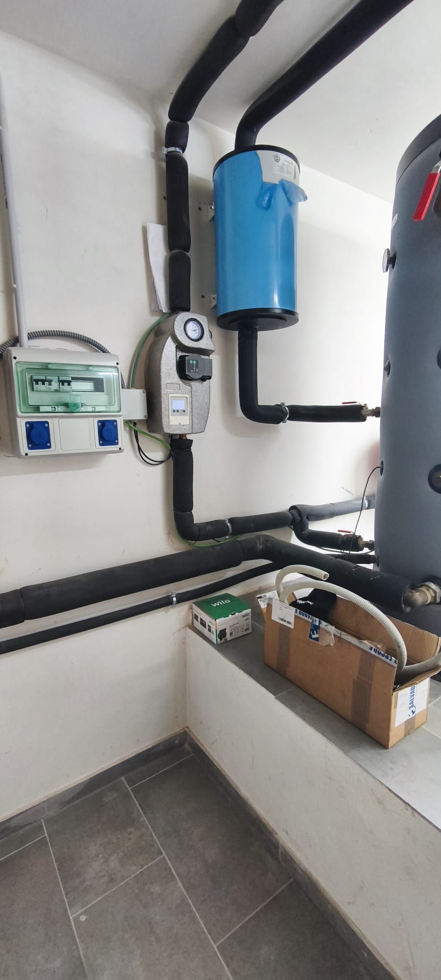 Rincón de servicios con contador eléctrico, depósito azul, tuberías y cajas de cartón en una habitación alicatada