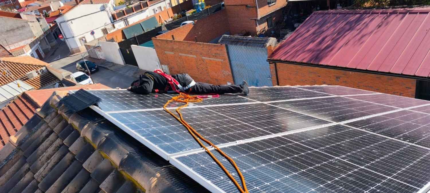 Trabajador instalando paneles solares en un tejado, con cuerdas de seguridad naranjas y edificios de ladrillo rojo cerca