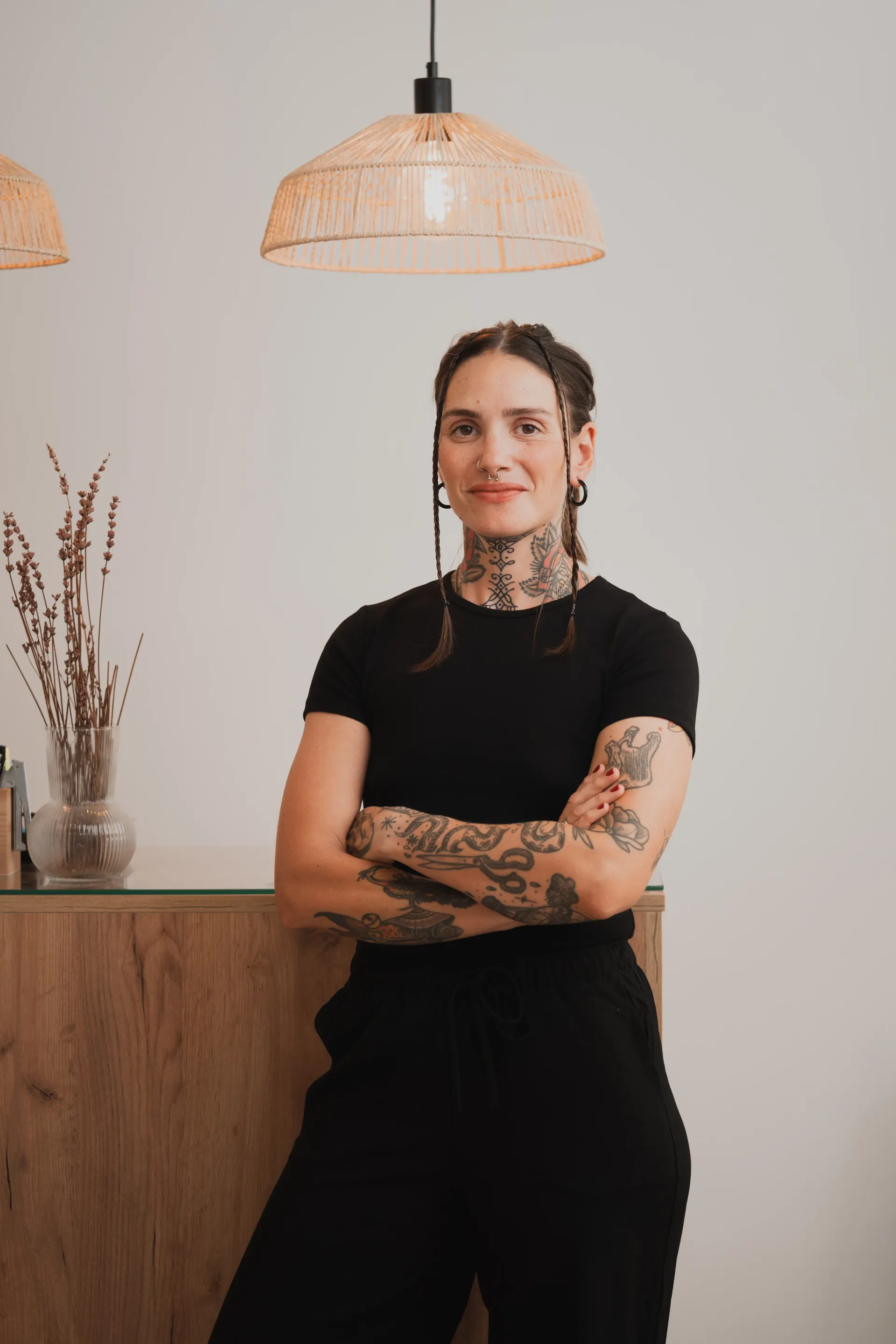 Mujer con tatuajes de pie, con los brazos cruzados, cerca de un mostrador de madera, bajo una lámpara de mimbre.