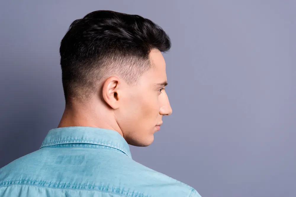 Hombre con pelo corto y oscuro, corte degradado, vistiendo una camisa azul verdosa, de perfil, sobre un fondo gris.