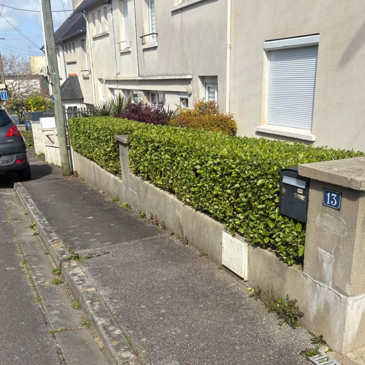 Trottoir résidentiel étroit longeant une maison, avec une haie, un mur de pierre, une voiture garée et le numéro 13.